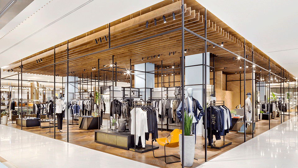 The menswear department is centered on a central pavilion, defined by a slatted oak ceiling raft and fleshed out with herringbone leather paneled walls, end-grain oak flooring, warm lacquers and dark metal frames.