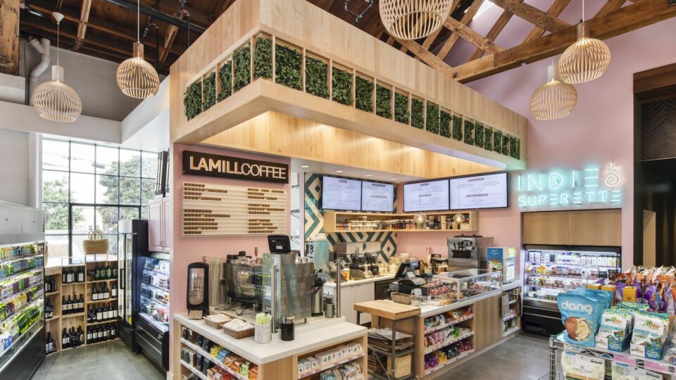 The LAMILLl Coffee counter features a sleek white Meganite countertop and natural light wood finishes. Digital display menus line the back and sides of the counter’s custom white and teal geometric tile wall. The service counter is topped with a drop-down soffit that is outfitted with lush planters, while handmade pressed birch wood pendant lights provide soft illumination. Hidden recessed lighting also assists in brightening the counter and the rest of the market.