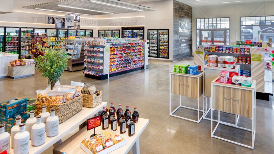 Nesting tables and branded specialty bins showcase new products and key merchandising stories. The Beer Cave and refrigerated coolers are highlighted by a soft glow from the perimeter signage, while a suspended wood ceiling plane features all the classic favorites below.