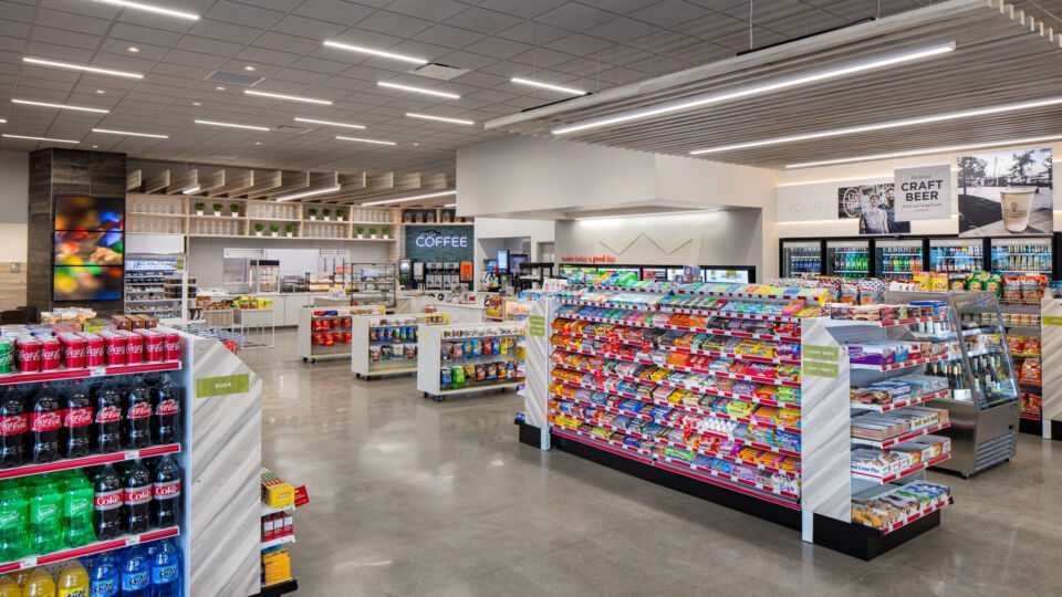 The main drive aisle leads shoppers into the store from the gas canopy, with open sightlines of the entire store. Digital marketing and a warm neon glow pull visitors back into the space. Linear lighting provides directional cues at varying heights and locations. Wood textured panels are incorporated on the endcaps.