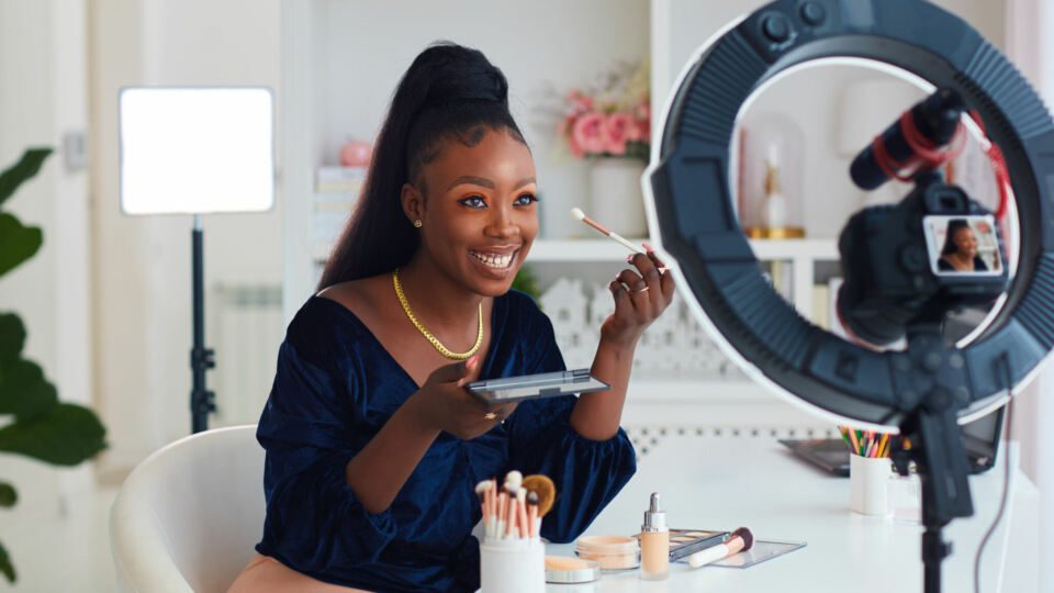 Happy African American woman putting on makeup during a live shopping tutorial.