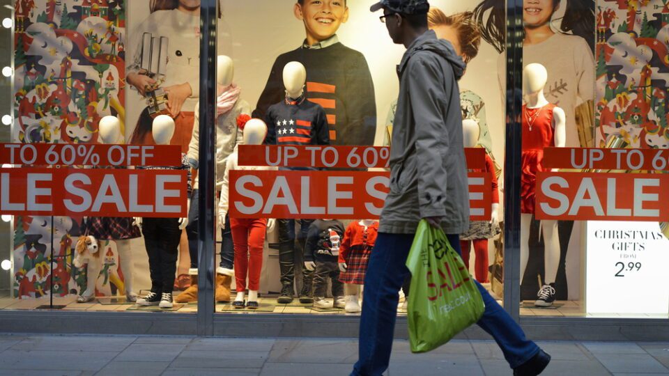 Man in hat and glasses walking past a retail store that has "sale" signs along the front of it