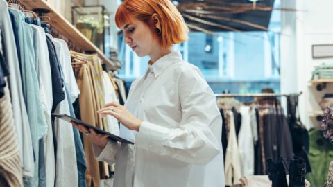 Young female associate with red hair and in a white shirt. She's using a tablet as she's consulting a rack of clothes.