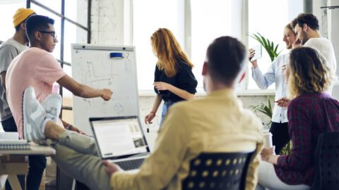 A group of young employees in a store design team sitting around an open office space talking and collaborating. Backs turned to the camera.