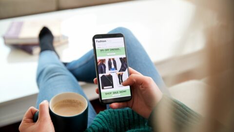 A woman sitting in a living room, feet up with a cup of coffee, interacting with an ecommerce site experience on her smartphone.