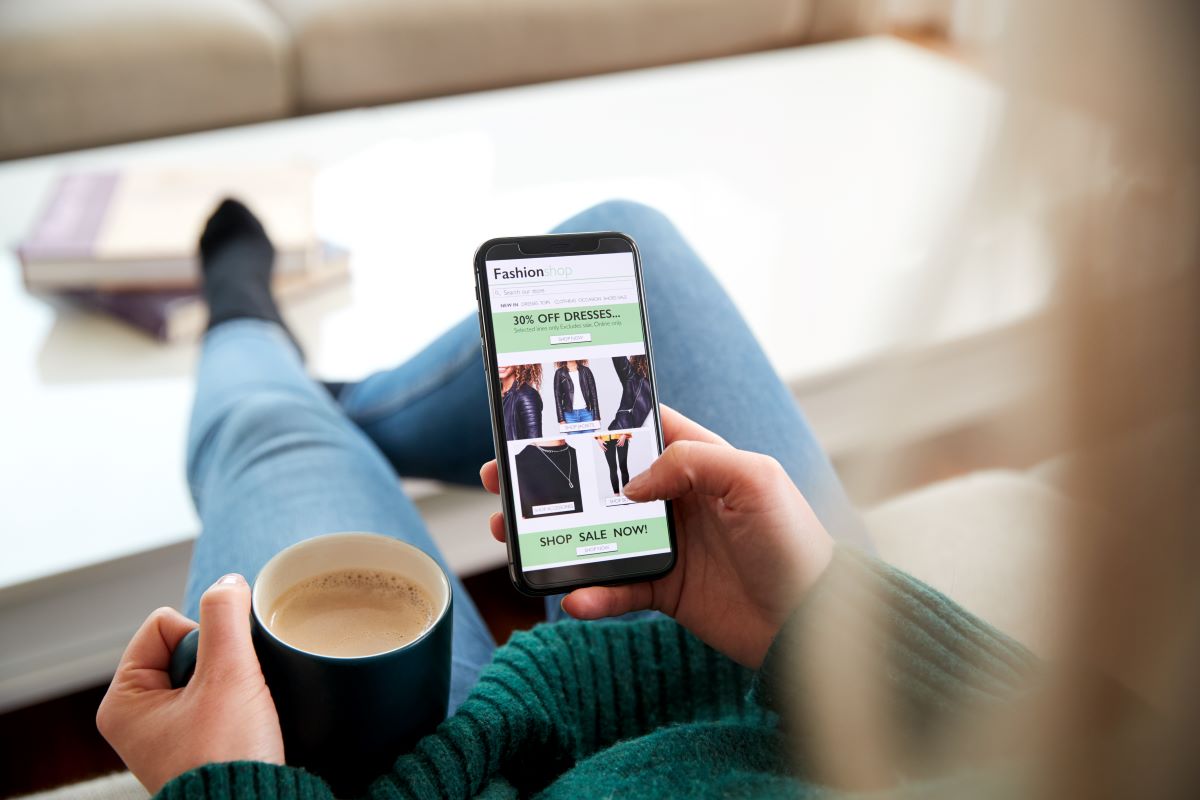 A woman sitting in a living room, feet up with a cup of coffee, interacting with an ecommerce site experience on her smartphone.